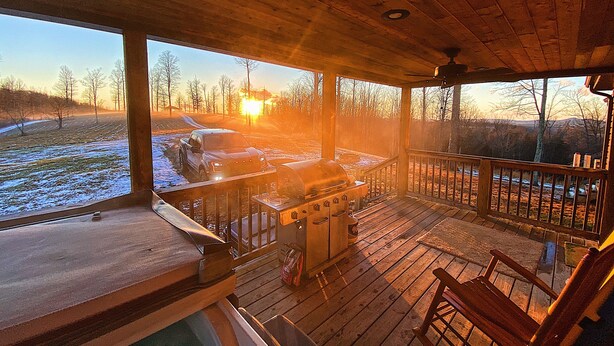 Una terraza de madera con una parrilla y una silla, con vista a un paisaje nevado que incluye un vehículo y árboles.