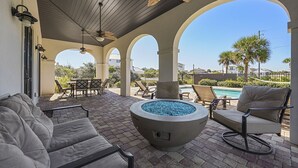 Eine Terrasse im Freien mit Feuerstelle, Sitzgelegenheiten und Blick auf Pool und Palmen.