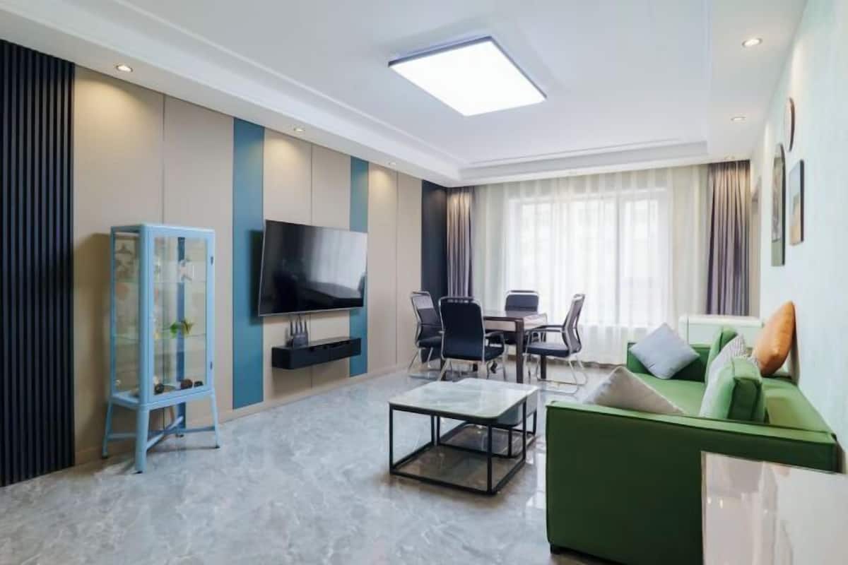 A modern living room with a flat-screen TV, a glass coffee table, a green sofa, and a dining area with chairs.