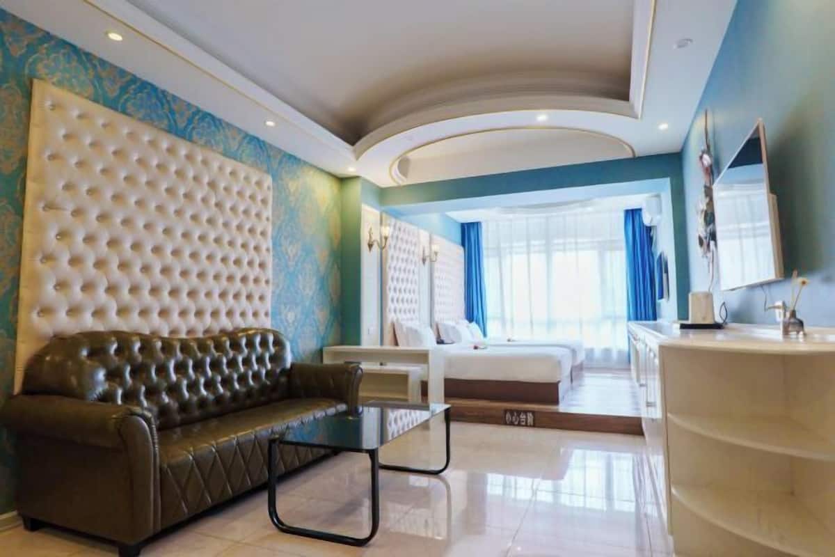 Una habitación de hotel moderna con cabecera capitonada, un sofá de cuero, una mesa de centro de vidrio, una cama con ropa blanca, un tocador con espejo y un ventanal con cortinas.