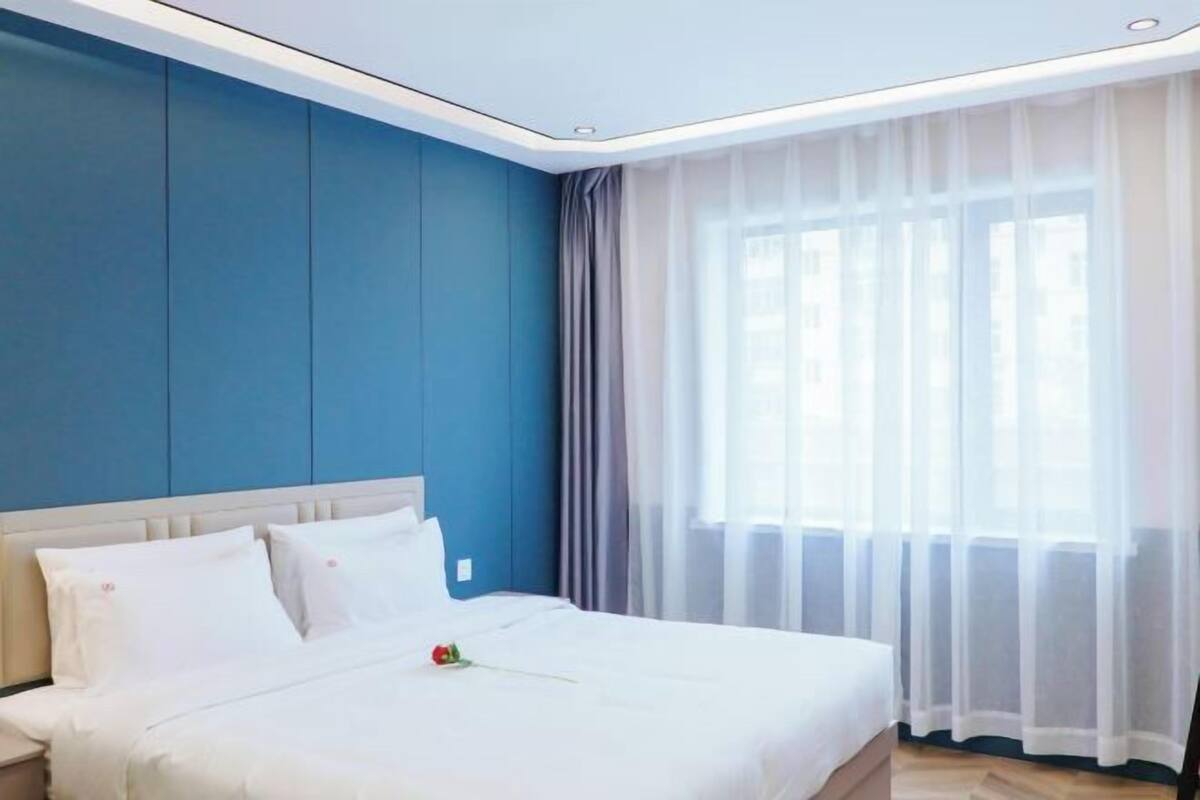 Habitación de hotel con cama, mesita de noche, ventana con cortinas translúcidas y pared azul.