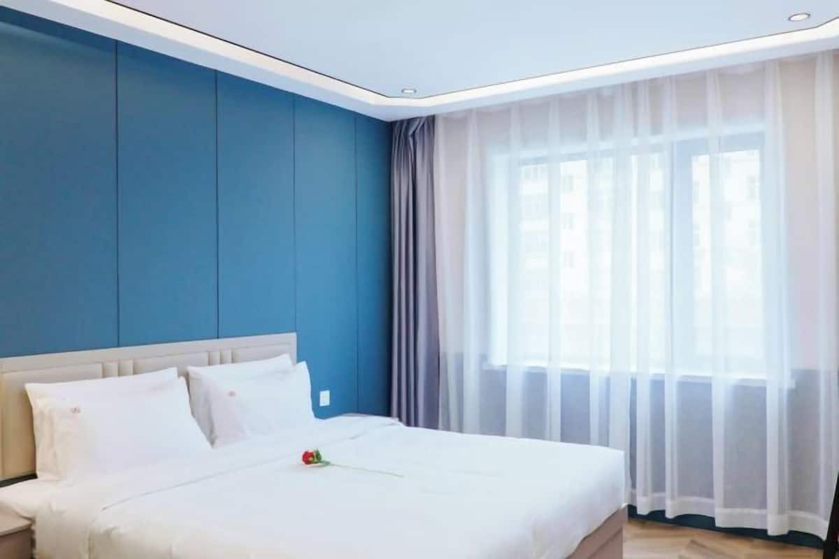 A hotel room with a bed, a nightstand, a window with sheer curtains, and a blue wall.