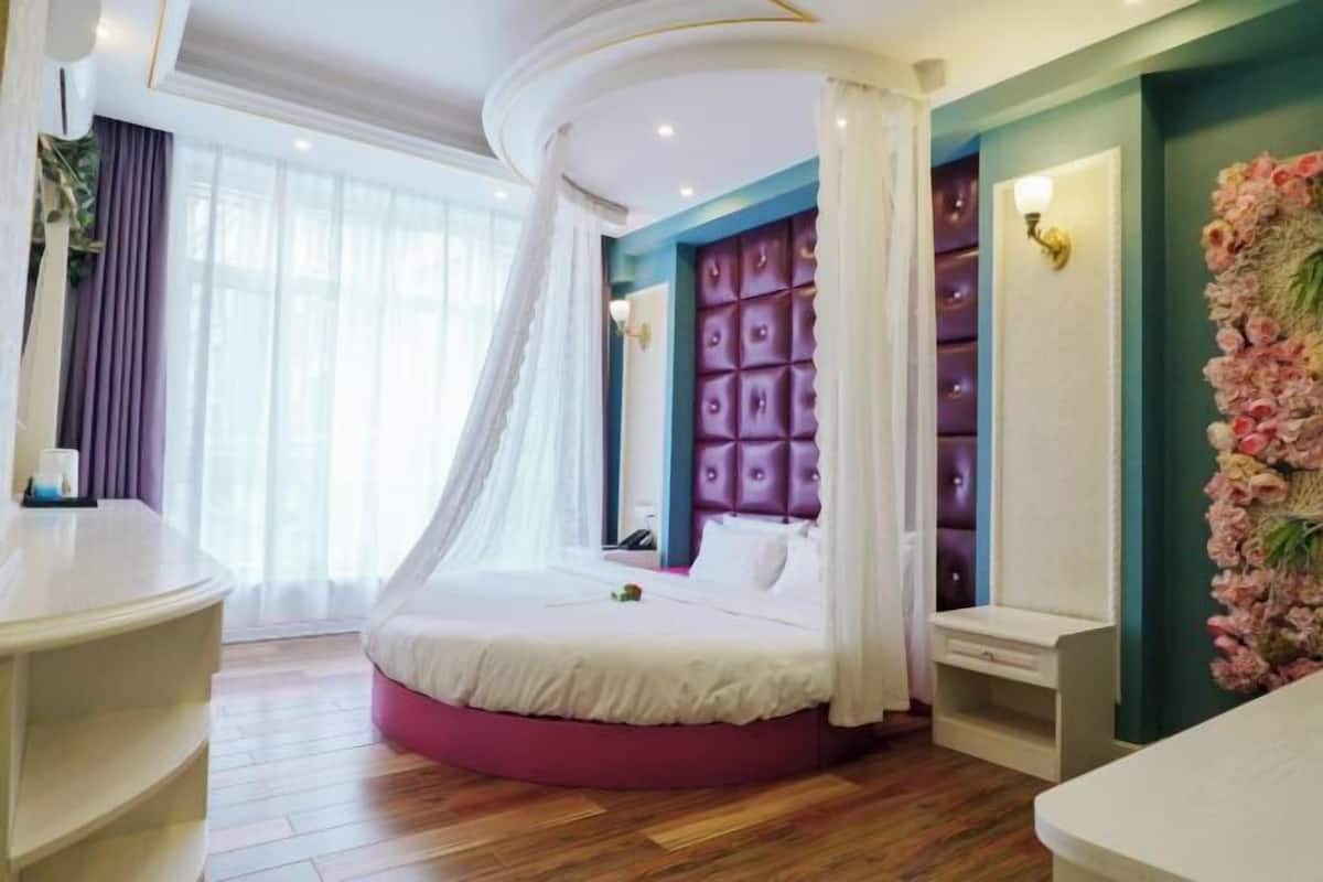 Un dormitorio con cama curvada, una mesita de noche, una lámpara fijada a la pared y un arreglo floral decorativo.