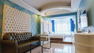 Una habitación de hotel moderna con una cabecera capitonada, un sofá de cuero, una mesa de centro de vidrio, una cama con ropa blanca y un espejo grande, y un baño con lavamanos blanco.