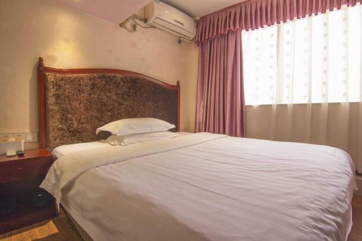 A hotel room with a large bed, a wooden headboard, a nightstand, an air conditioning unit, and a window with curtains.