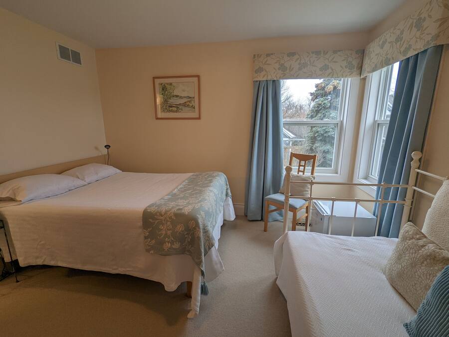 A bedroom with two beds, a chair, a window with curtains, a radiator, and a framed picture on the wall.