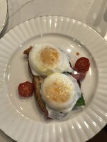 Un plato con dos huevos pochados sobre tostada, una rodaja de tomate y una loncha de jamón.