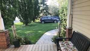 A wooden deck with a bench, a brick wall, and a view of a car and trees in the background. - Dicksons Family Suite B&B (Niagara-on-the-Lake)