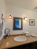 Un baño con un lavamanos blanco, un espejo redondo, un perchero, dos luces empotradas en la pared, un espejo enmarcado y una imagen en la pared.