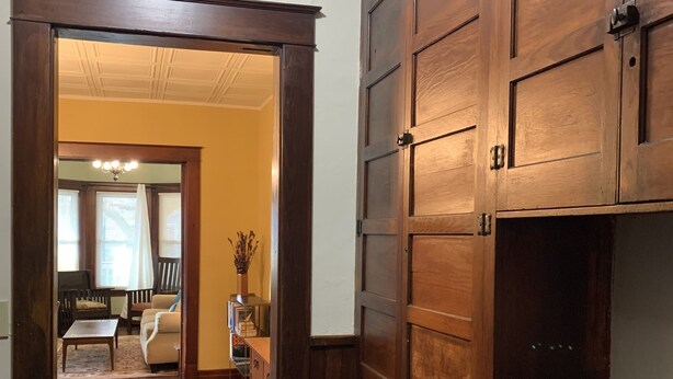 A hallway with wooden flooring and a large wooden door. There is a dining area visible in the background with a chandelier and furniture. - The Red Brick Inn - 10 Minute Walk to the Falls (Niagara Falls)