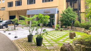A building with a beige facade, multiple windows, and a sign with Japanese characters. There are potted plants and a stone pathway in front.