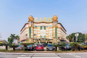 A multi-story hotel with a central entrance, a parking lot with several cars, and palm trees in the foreground. - The Dynast's Hotel (Foshan)