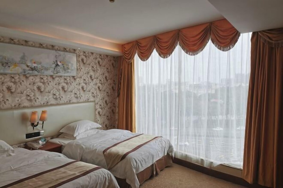 Een hotelkamer met twee bedden, een groot raam met dunne gordijnen, een schilderij aan de muur en een hoofdbord met een bloemenprint.