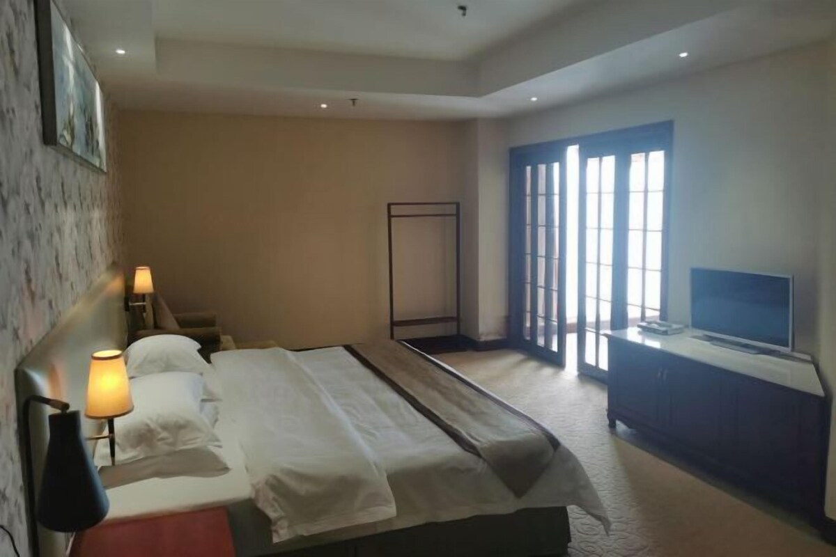 Een hotelkamer met een bed, bedlampen, een televisie en een groot raam met jaloezieën.