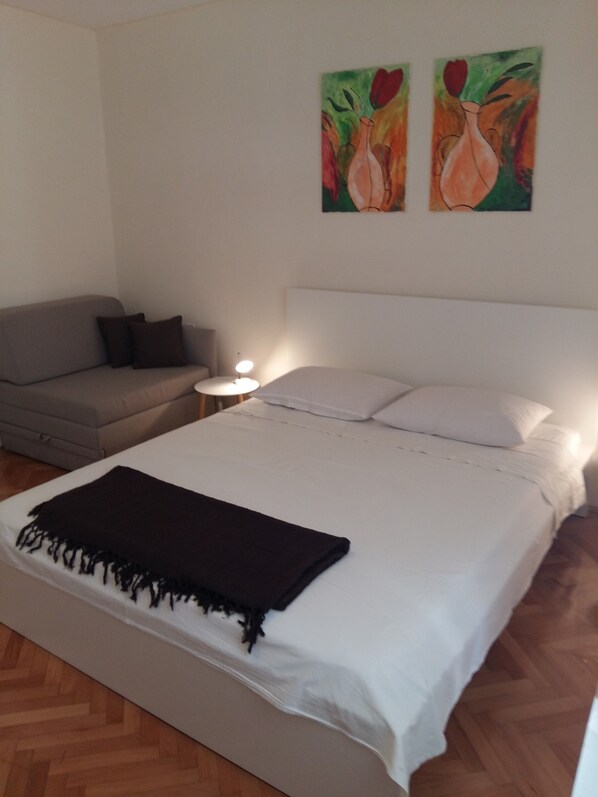 Um quarto com cama, mesa de cabeceira, abajur, sofá e duas pinturas na parede.