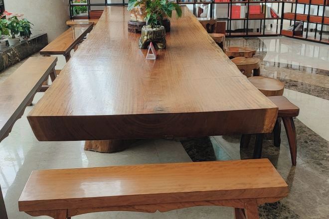 A long wooden table with benches, surrounded by bookshelves and potted plants in a well-lit indoor space.