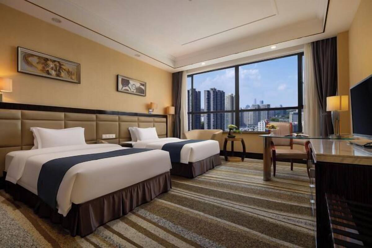 Habitación de hotel con dos camas, un escritorio, una silla y un ventanal con vista a la ciudad.