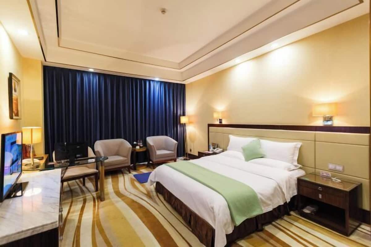 Habitación de hotel con cama, dos sillones, un escritorio con computadora, televisor y un gran ventanal con cortinas.