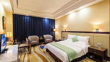 Habitación de hotel con cama, dos sillones, un escritorio con computadora, televisor y un gran ventanal con cortinas.