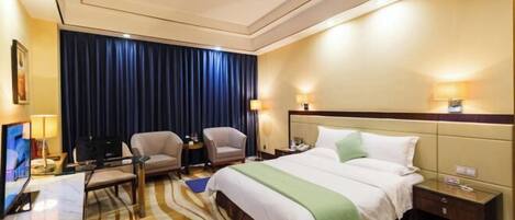 A hotel room with a bed, two armchairs, a desk with a computer, a TV, and a large window with curtains.