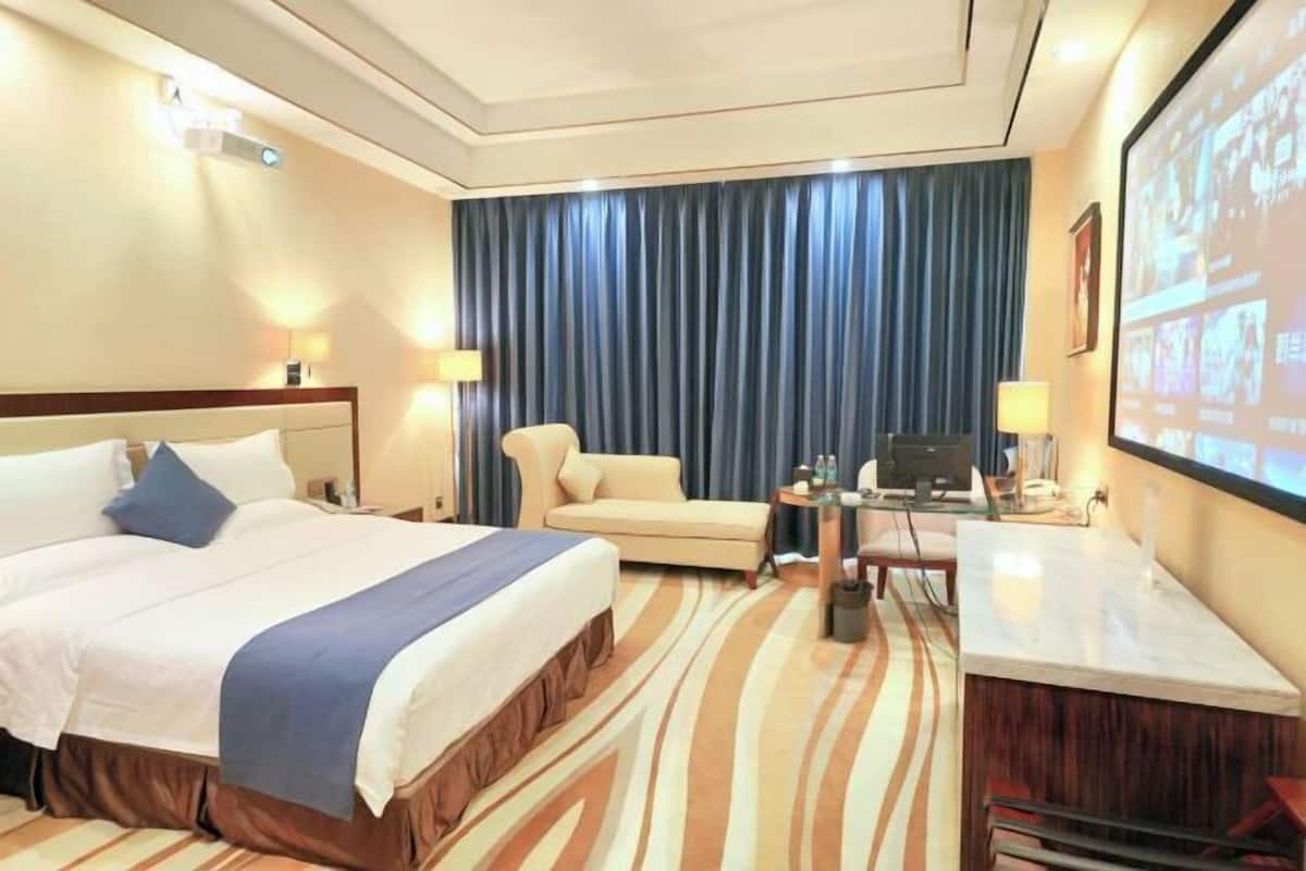 Habitación de hotel con una cama grande, un sofá, un escritorio con computadora y una pantalla de televisión mostrando imágenes.