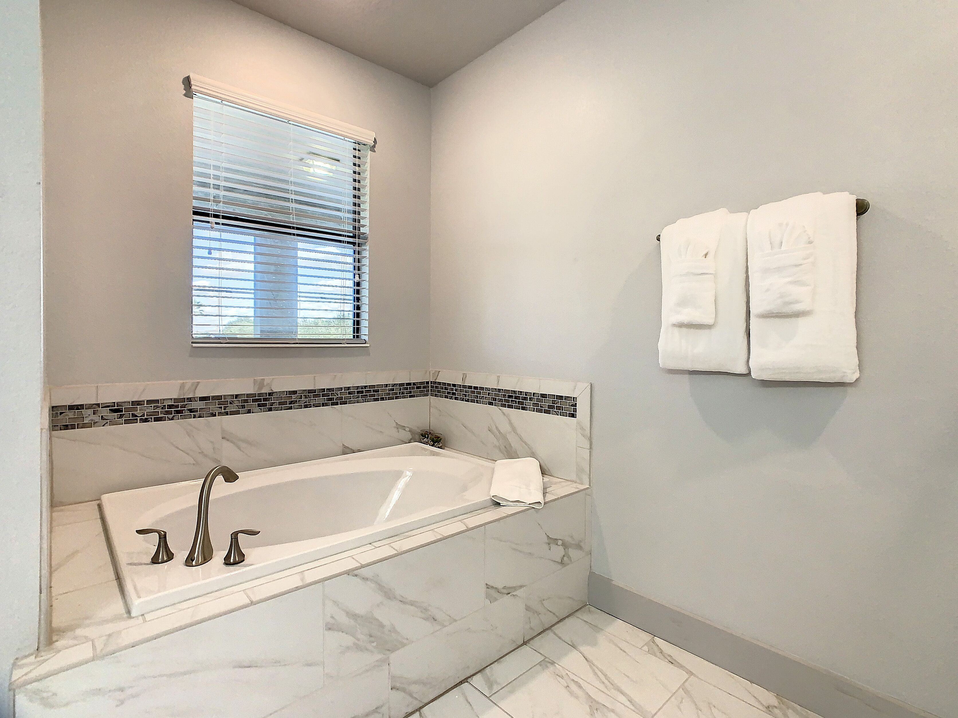 A bathroom with a marble bathtub, a window with blinds, and two towels hanging on a rack.