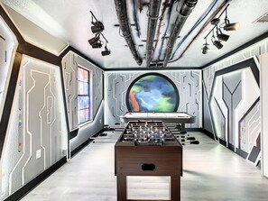 A room with a foosball table, a large window with a view of space, and a wall with a space-themed design.