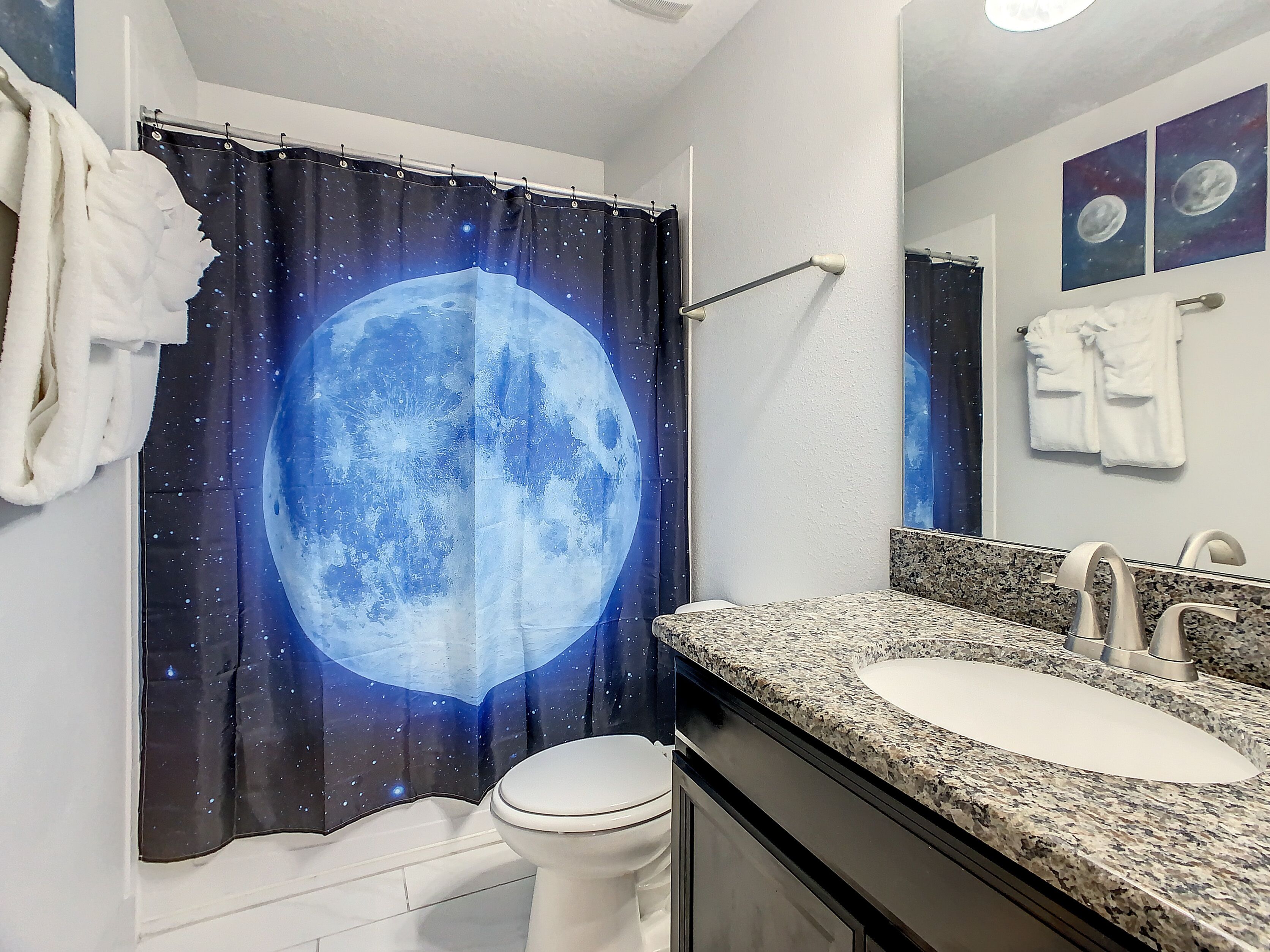 Um banheiro com cortina de chuveiro temática de lua, vaso sanitário branco e pia com bancada de granito e espelho acima.