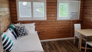 A small cabin room with a wooden interior, a bed with patterned pillows, a table with two chairs, and a window with an air conditioning unit. - Studio across the street from beach (Hauula)