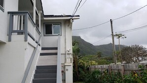 A residential building with a staircase leading to a door, surrounded by lush greenery and a mountainous backdrop. - Studio across the street from beach (Hauula)