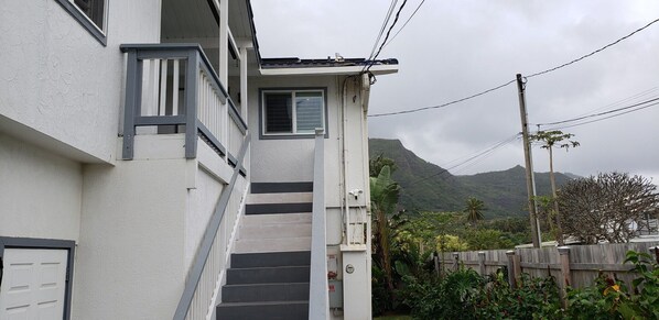 A residential building with a staircase leading to a door, surrounded by lush greenery and a mountainous backdrop.