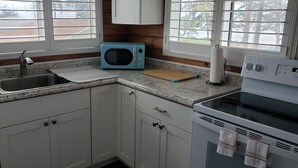 A kitchen with white cabinets, a blue microwave, a sink, a cutting board, and a stove. - Studio across the street from beach (Hauula)