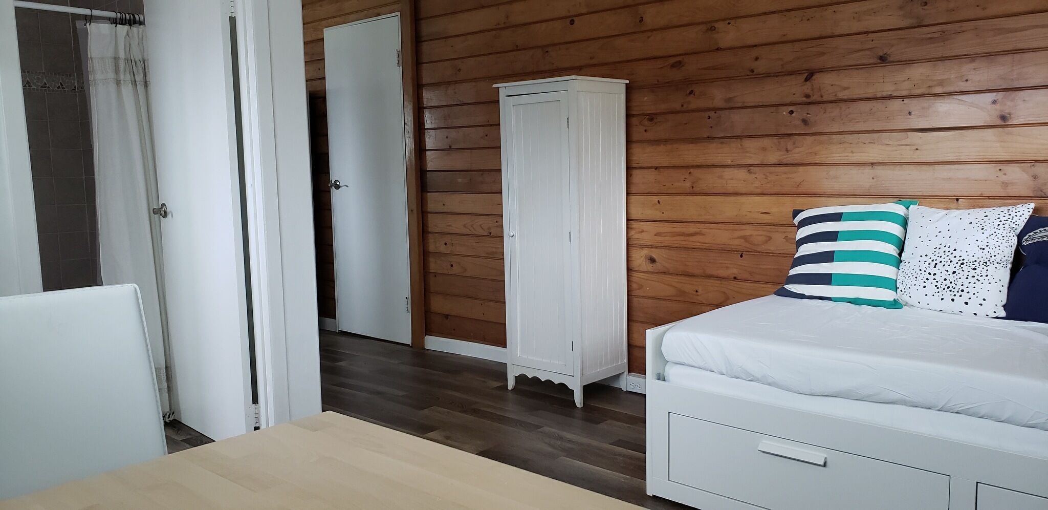 Een slaapkamer met een houten vloer, een witte kledingkast, een bed met gestreepte kussens en een witte deur.