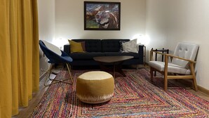 A living room with a dark blue sofa, a round ottoman, a wooden chair, and a colorful woven rug. - Life at the Dairy Farm (New Enterprise)