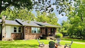 Une maison à deux étages, avec une façade en briques et un porche couvert. On y trouve deux chaises de jardin et une petite table sur la pelouse devant la maison.