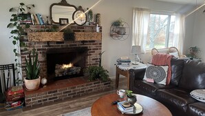 Un salon chaleureux avec une cheminée en briques, un canapé en cuir et une table basse ornée de livres et d’une plante.