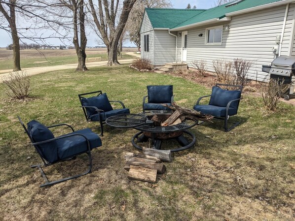 Eine Terrasse mit vier blauen Stühlen, einem runden Tisch und einer Feuerstelle mit Holzscheiten.