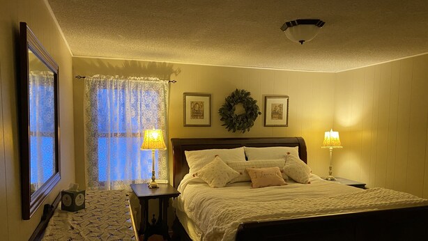 A bedroom with a bed, two bedside tables with lamps, a nightstand, a decorative wreath, and a mirror on the wall.