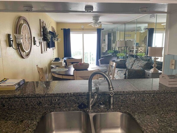 A modern kitchen with granite countertops, stainless steel sinks, and a dining area with a round table and chairs. - Family Friendly Condo in Oceanfront Building (North Myrtle Beach)