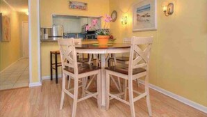 Zona pranzo con tavolo e sedie in legno, cucina sullo sfondo e parete gialla con orologio e quadro incorniciato.