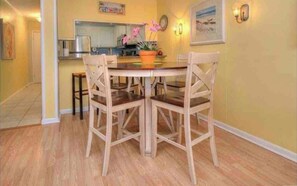 A dining area with a wooden table and chairs, a kitchen in the background, and a yellow wall with a clock and a framed picture.