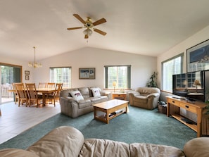A spacious living room with a high ceiling, a ceiling fan, a dining area with wooden furniture, and large windows providing natural light.