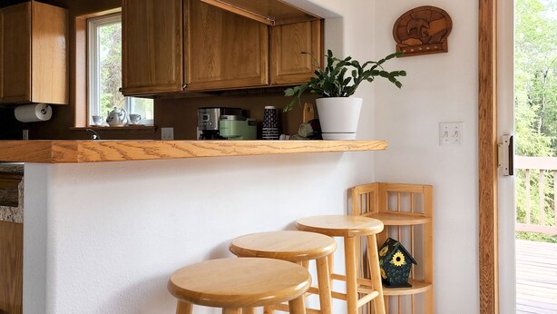 A kitchen with wooden cabinets, a white countertop, and a potted plant. There are three wooden stools and a wooden chair. - Alaska Fly In / Drive In, Kenai Peninsula Lodging (Sterling)