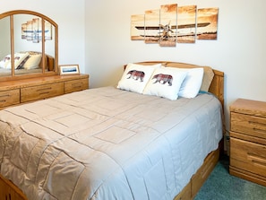 A bedroom with a large bed, wooden headboard, a dresser, a mirror, and a wall with a multi-panel artwork.