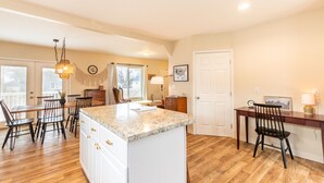 A spacious kitchen with a central island, wooden dining table, and chairs. There is a wooden desk with a chair and a lamp. - Kid-Friendly 2 Bedroom close to Idaho Center & NNU (Nampa)