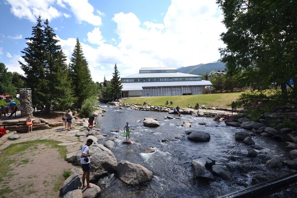 A river with rocks and people standing on them. There is a building in the background with a flat roof. - Downtown Location walk to Main Street and Gondola from this updated Condo (Breckenridge)