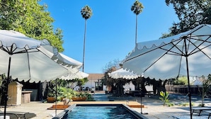 A swimming pool with two white umbrellas, surrounded by potted plants and a clear sky.