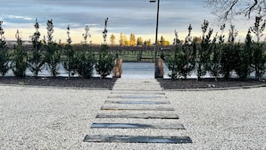 A gravel pathway leading to a bridge over a body of water, surrounded by trees and a fence.