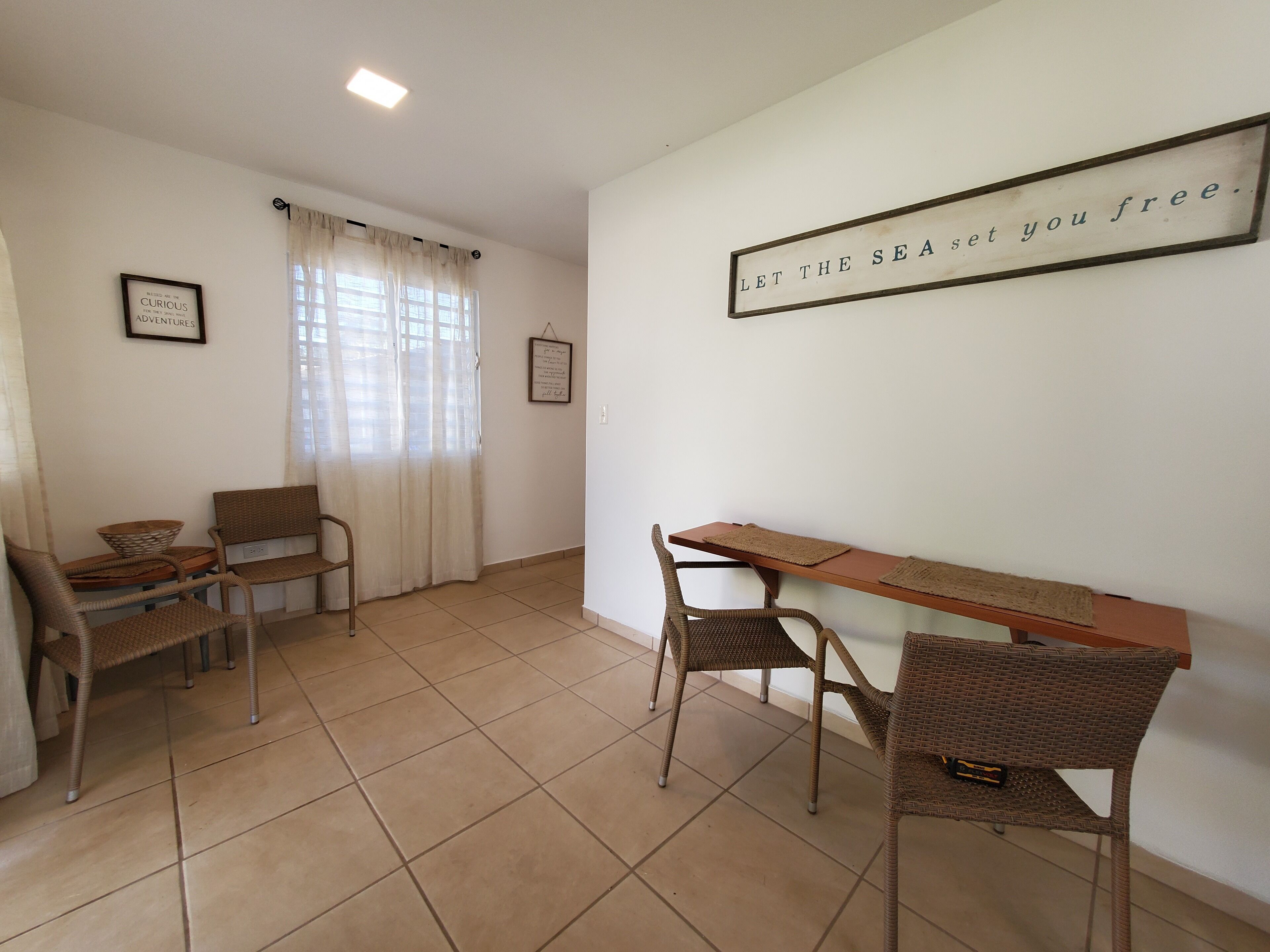 Una habitación con una mesa de comedor, sillas, un ventanal con cortinas y un cartel en la pared.