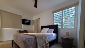A bedroom with a bed, a TV mounted on the wall, a window with blinds, and a small trash bin. - Comfy apartment 8 minute walk from beach (Aguadilla)